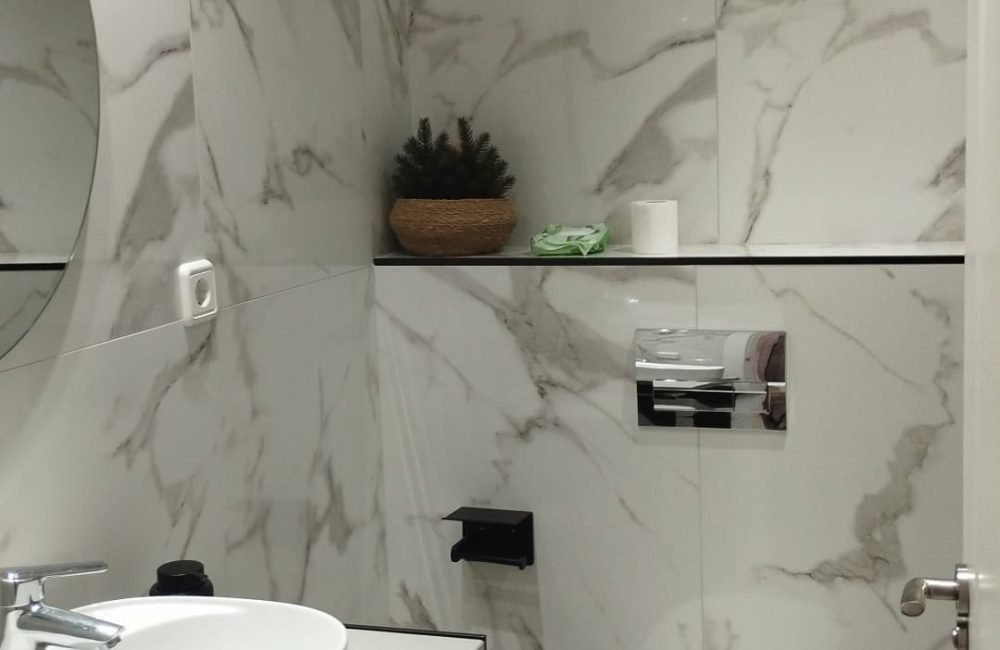 Baño moderno con paredes de mármol blanco, lavabo blanco sobre una encimera, espejo redondo y un inodoro blanco. Un estante sobre el inodoro tiene algunos objetos decorativos.